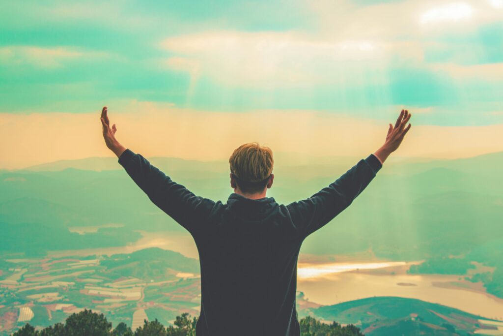 A man stands with arms raised, embracing the vast landscape from a mountain viewpoint.
