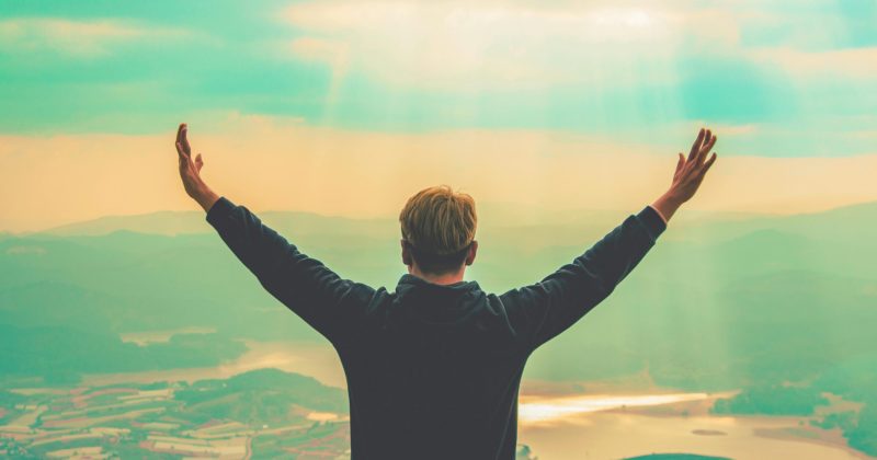 A man stands with arms raised, embracing the vast landscape from a mountain viewpoint.