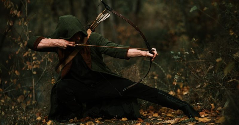 Medieval archer crouched in forest, aiming a bow with precision. Autumn foliage surrounds.